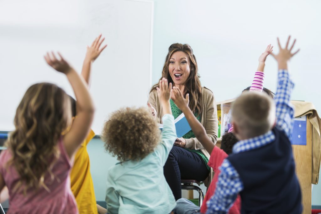 A female, Asian preschool or kindergarten teacher in the classroom, sitting in front of a group of multi-ethnic children, reading a book. The boys and girls are raising their hands to answer a question. There is a large, blank whiteboard in the background for copy.